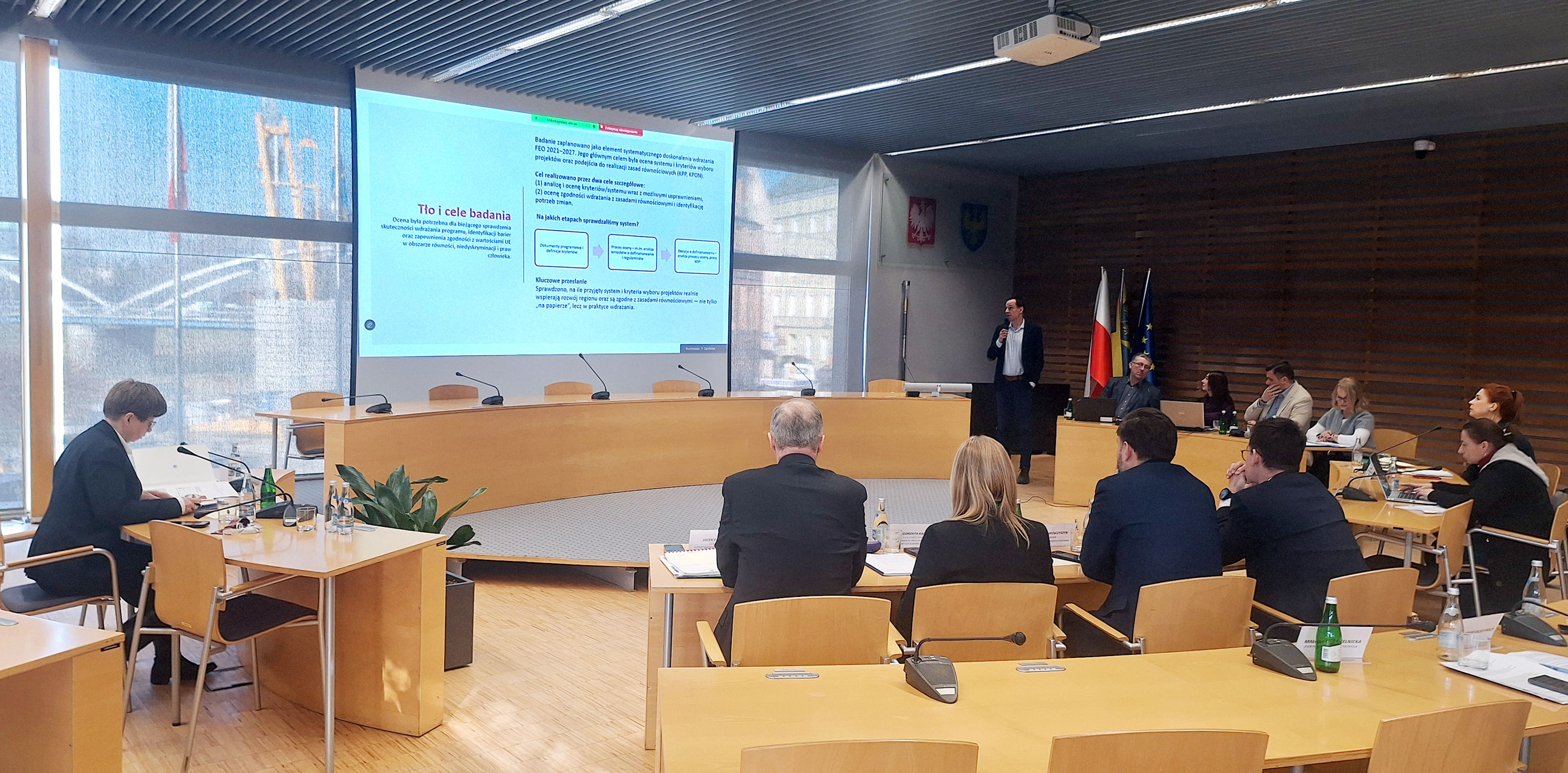 Widok ogólny sali obrad podczas posiedzenia Komitetu Monitorującego Fundusze Europejskie dla Opolskiego 2021–2027 w Urzędzie Marszałkowskim Województwa Opolskiego. Członkowie Komitetu uczestniczą w obradach, a na ekranie wyświetlana jest prezentacja dotycząca celów badania ewaluacyjnego.