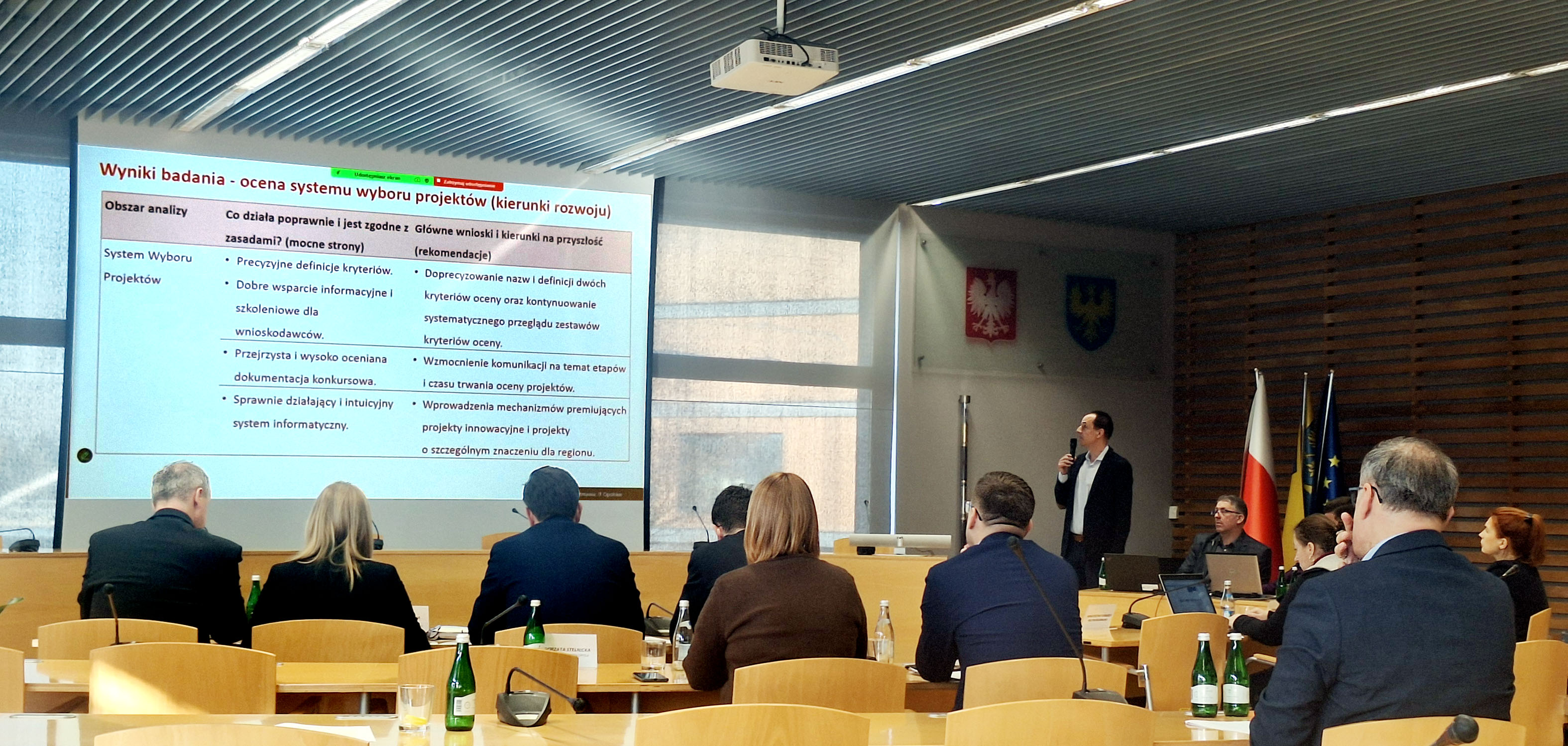 Wystąpienie przedstawiciela Wykonawcy podczas prezentacji wyników badania ewaluacyjnego na posiedzeniu Komitetu Monitorującego FEO 2021–2027. Prelegent omawia slajd dotyczący oceny systemu wyboru projektów, a uczestnicy słuchają prezentacji.