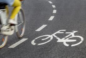 Asfaltowa droga rowerowa, z namalowanym białym rowerem. Po lewej stronie poza ostrością jadąca na rowerze osoba w żółtych spodniach - widać ją do połowy. 