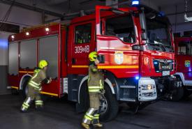 Dwóch strażaków w ubraniach ochronnych wsiada do czerwonego wozu strażackiego stojącego w garażu remizy