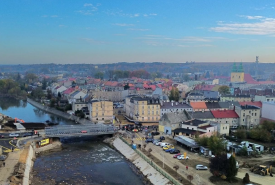 Widok z lotu ptaka na miasto w powiecie nyskim. Na pierwszym planie widoczny jest most tymczasowy nad rzeką, wybudowany po powodzi. W tle znajduje się zabudowa miejska i kościół z dwiema wieżami.