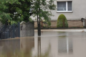 Zalany wodą fragment ulicy przed budynkami mieszkalnymi. Woda sięga do bram posesji i ogrodzeń, a nawierzchnia drogi jest całkowicie pokryta wodą. Zdjęcie dokumentuje skutki powodzi.