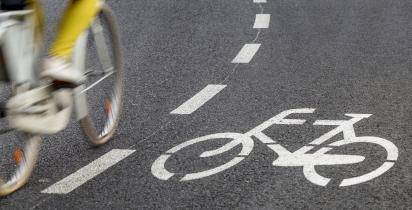 Asfaltowa droga rowerowa, z namalowanym białym rowerem. Po lewej stronie poza ostrością jadąca na rowerze osoba w żółtych spodniach - widać ją do połowy. 