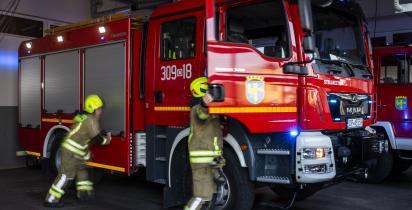 Dwóch strażaków w ubraniach ochronnych wsiada do czerwonego wozu strażackiego stojącego w garażu remizy