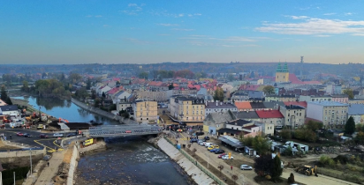Widok z lotu ptaka na miasto w powiecie nyskim. Na pierwszym planie widoczny jest most tymczasowy nad rzeką, wybudowany po powodzi. W tle znajduje się zabudowa miejska i kościół z dwiema wieżami.