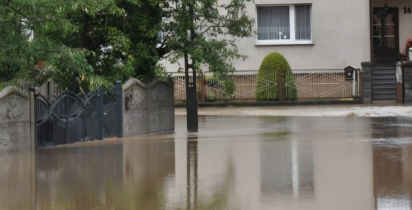 Zalany wodą fragment ulicy przed budynkami mieszkalnymi. Woda sięga do bram posesji i ogrodzeń, a nawierzchnia drogi jest całkowicie pokryta wodą. Zdjęcie dokumentuje skutki powodzi.