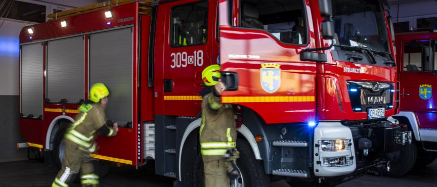 Dwóch strażaków w ubraniach ochronnych wsiada do czerwonego wozu strażackiego stojącego w garażu remizy