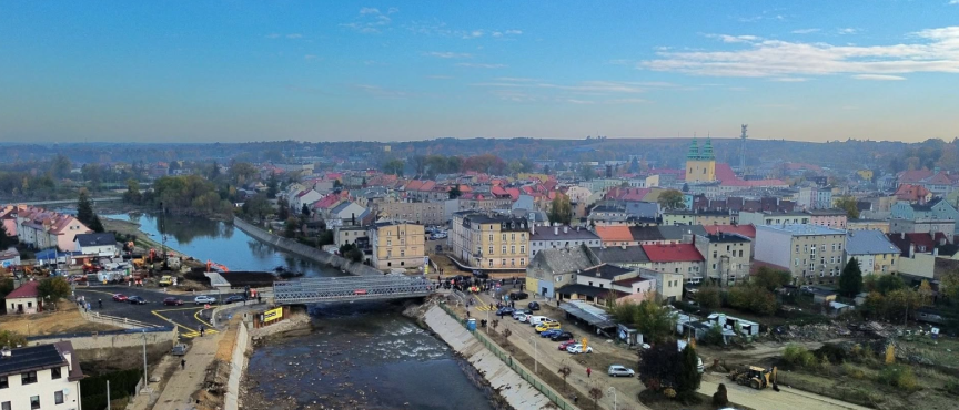 Widok z lotu ptaka na miasto w powiecie nyskim. Na pierwszym planie widoczny jest most tymczasowy nad rzeką, wybudowany po powodzi. W tle znajduje się zabudowa miejska i kościół z dwiema wieżami.