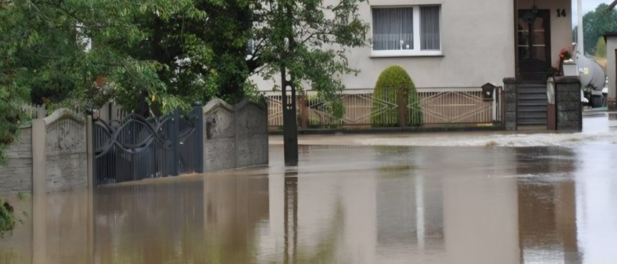 Zalany wodą fragment ulicy przed budynkami mieszkalnymi. Woda sięga do bram posesji i ogrodzeń, a nawierzchnia drogi jest całkowicie pokryta wodą. Zdjęcie dokumentuje skutki powodzi.