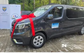 Samochód dostosowany do transportu osób potrzebujących wsparcia, udekorowany czerwoną wstążką, zaparkowany przed budynkiem. Obok znajduje się baner Regionalnego Ośrodka Polityki Społecznej w Opolu.