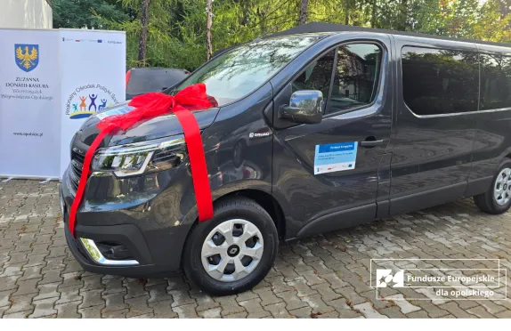 Samochód dostosowany do transportu osób potrzebujących wsparcia, udekorowany czerwoną wstążką, zaparkowany przed budynkiem. Obok znajduje się baner Regionalnego Ośrodka Polityki Społecznej w Opolu.